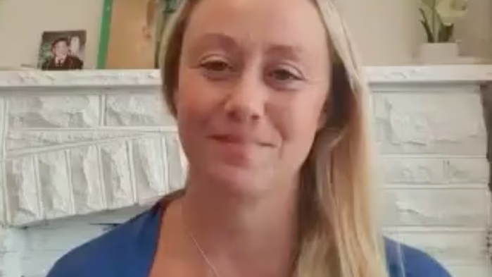 A woman with long blonde hair smiles softly while seated indoors. She is wearing a blue top. The background shows a white brick fireplace and some home decorations, including a plant and a framed photo. The image appears to be part of an online testimonial or video call, possibly related to yoga teacher training.