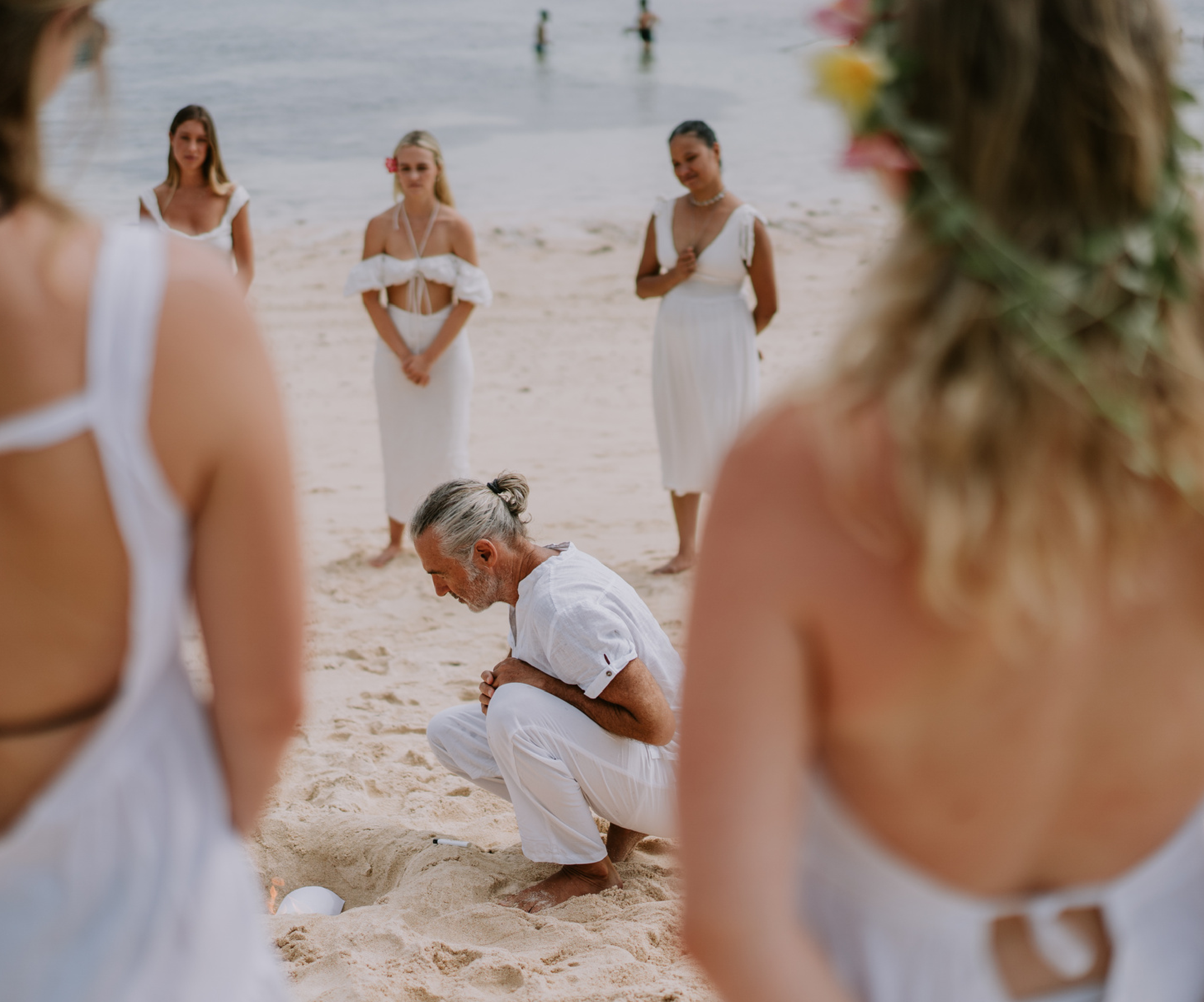 A serene beach ceremony with a group of individuals dressed in white, standing in a circle, as a teacher kneels in the sand. This setting embodies the essence of unforgettable and rejuvenating learning experiences in yoga teacher training, promoting a sense of community and spiritual connection.