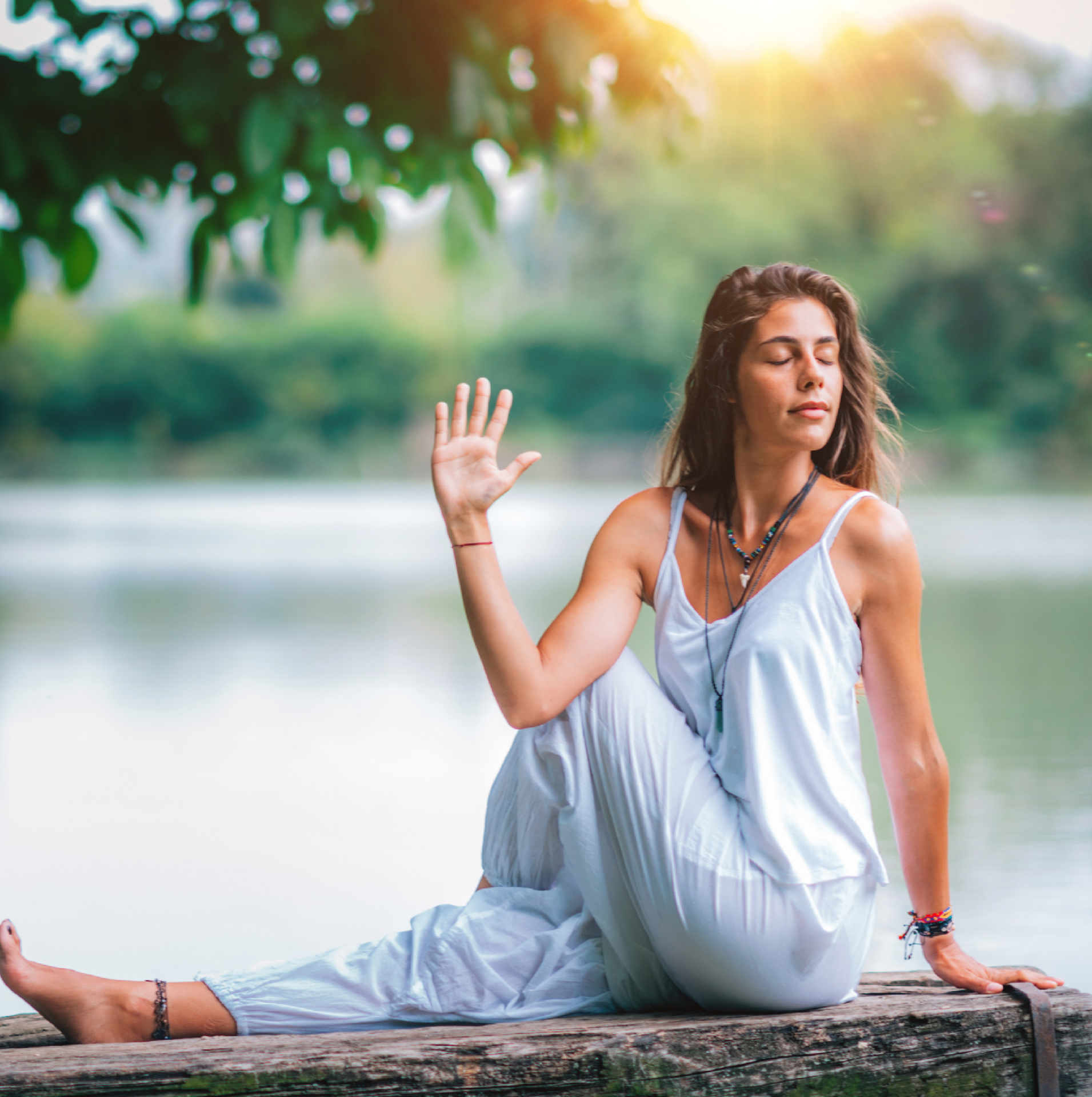 A woman practicing yoga outdoors by a peaceful lake, seated in a twist pose with her eyes closed, bathed in warm sunlight. The image evokes a sense of harmony and well-being, ideal for promoting an Online RYT-200 Yoga Teacher Training.