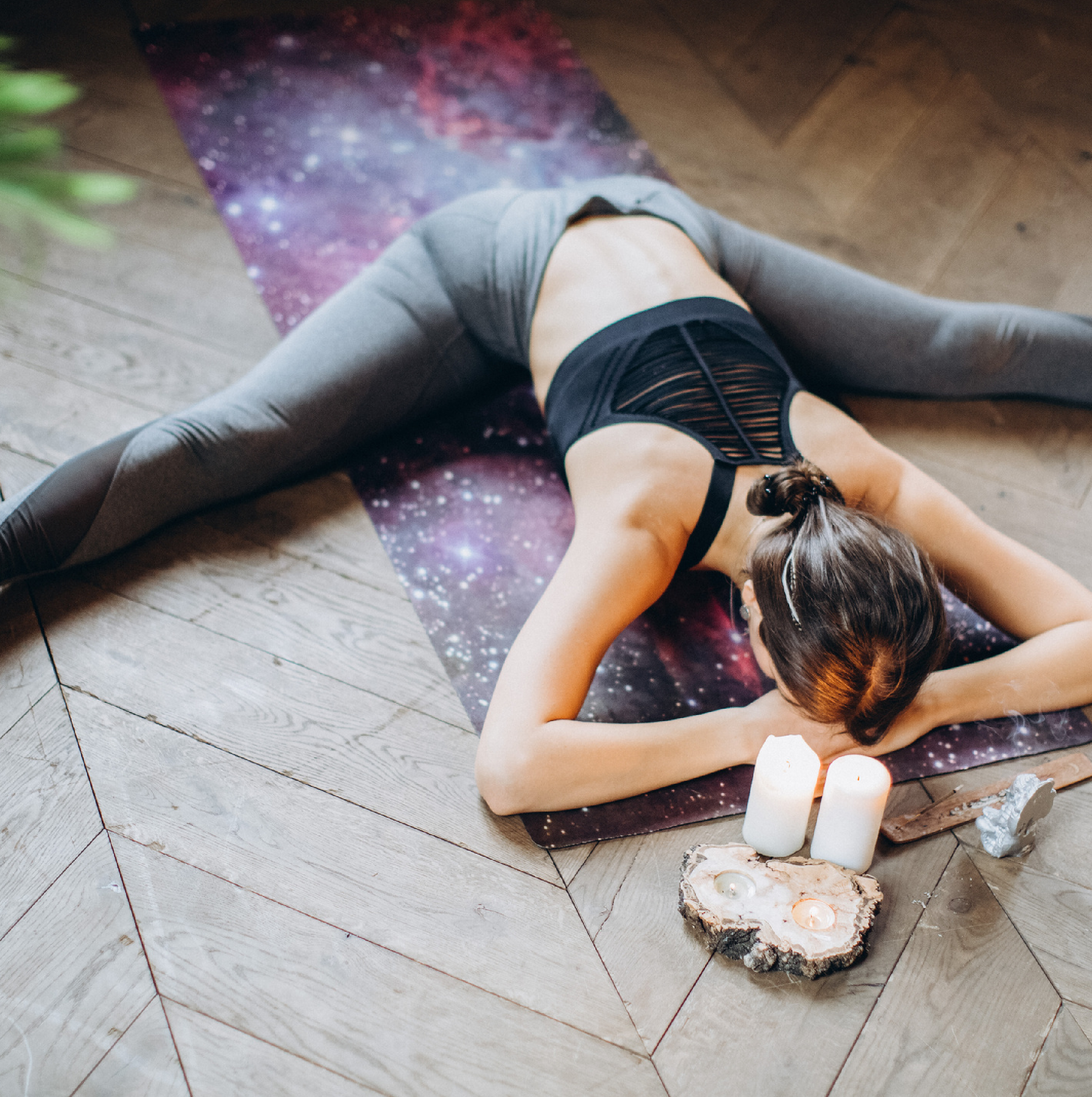 A woman in a wide-legged forward fold yoga pose on a cosmic-themed mat, with candles and crystals placed nearby, creating a calming and meditative atmosphere. The indoor setting and focus on relaxation make this image ideal for promoting an Online RYT-200 Yoga Teacher Training program.