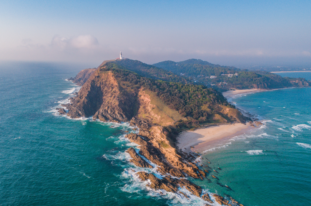 Aerial view of Byron Bay's iconic lighthouse perched on the edge of a rocky coastal cliff, surrounded by lush greenery and the clear blue ocean, showcasing the serene environment perfect for yoga teacher training.