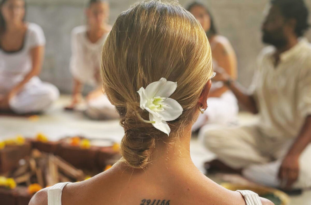 Close-up of a woman with a flower tucked into her hair, seated in a serene group setting during a yoga teacher training session in Sri Lanka, surrounded by people wearing white and calm natural tones.