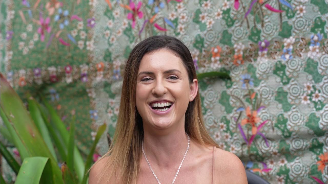 Jade smiling brightly while sharing her Santosha Yoga Teacher Training experience, seated in front of a vibrant green and floral fabric backdrop