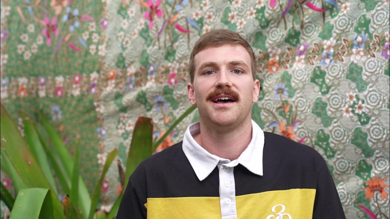 Lewis speaking in front of a colourful floral tapestry with tropical plants in the foreground