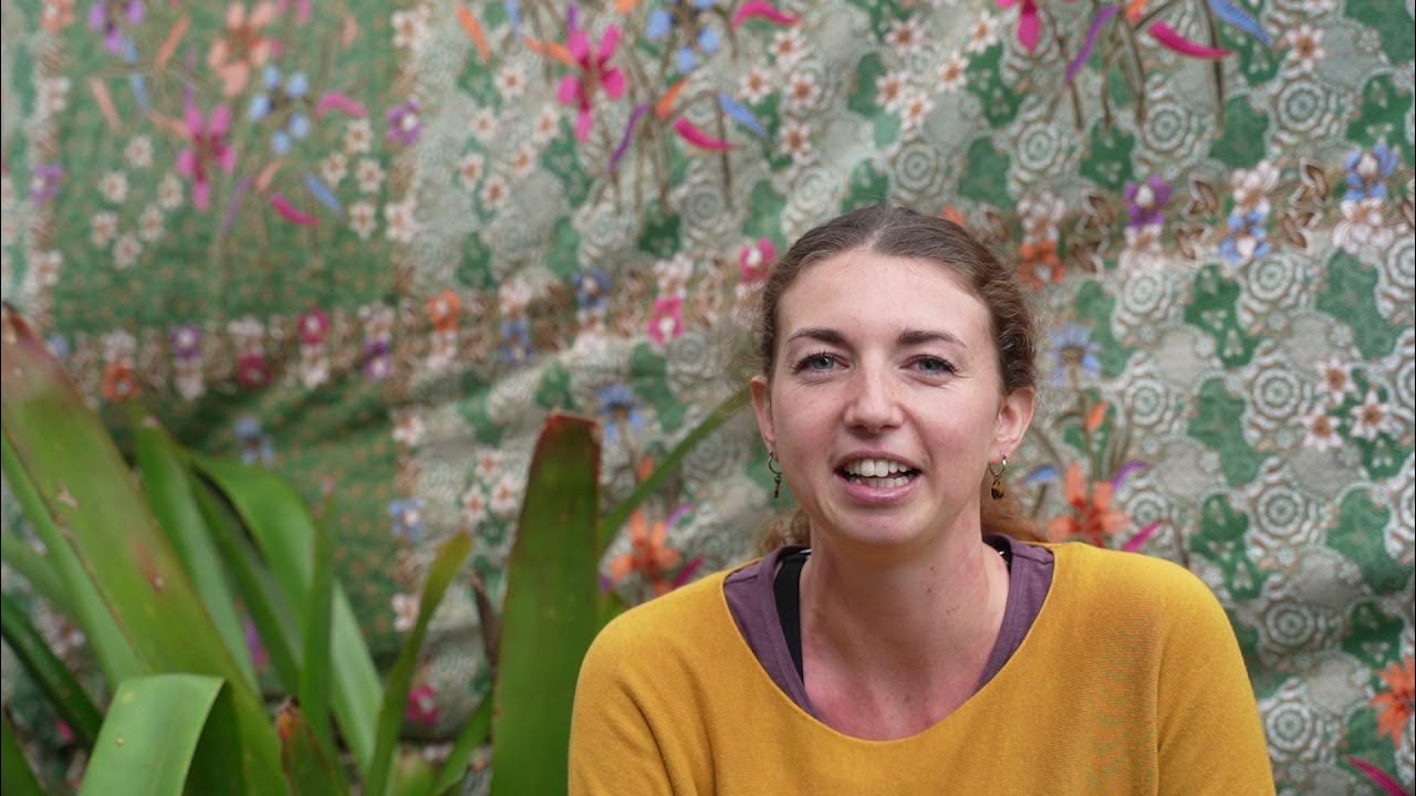 Roxie smiling and speaking during her yoga teacher training interview, with a green floral backdrop and tropical plants