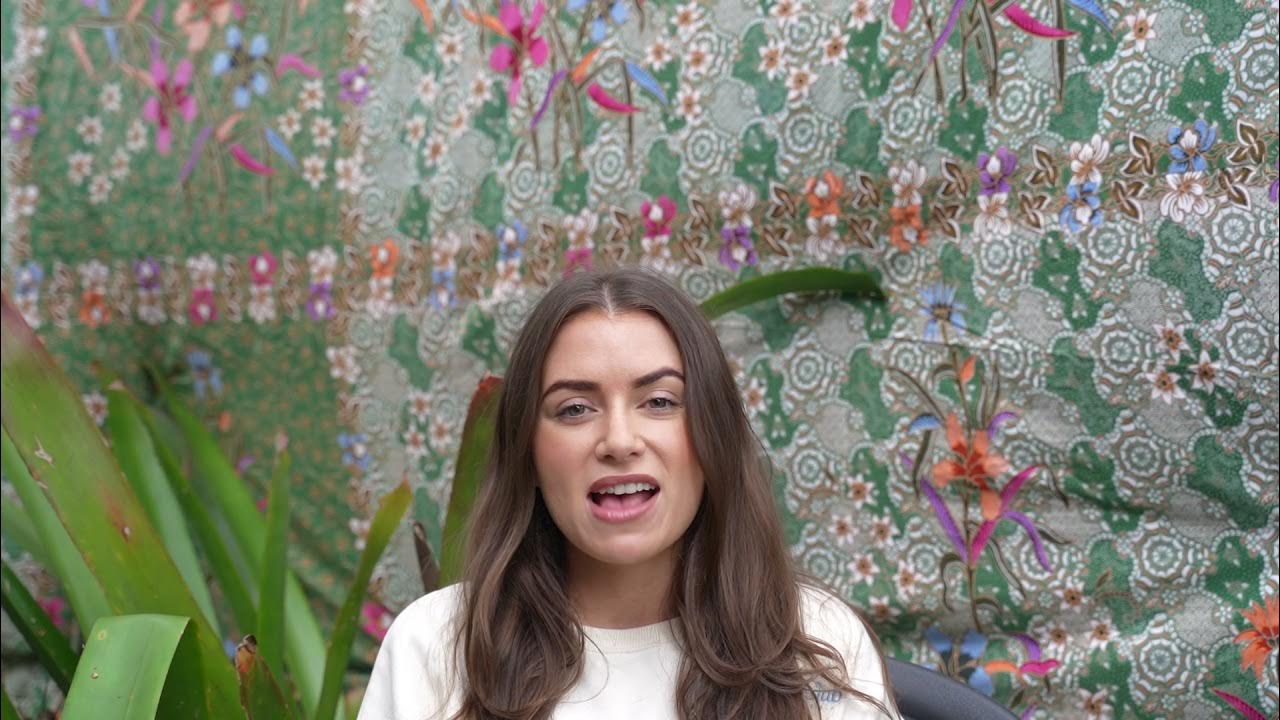 Sophia sharing her testimonial during Santosha Yoga Teacher Training, seated in front of a green floral-patterned backdrop with tropical plants