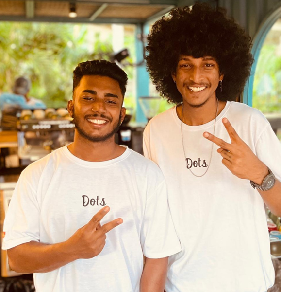 Two smiling men wearing 'Dots' shirts, making peace signs at a local Sri Lanka café