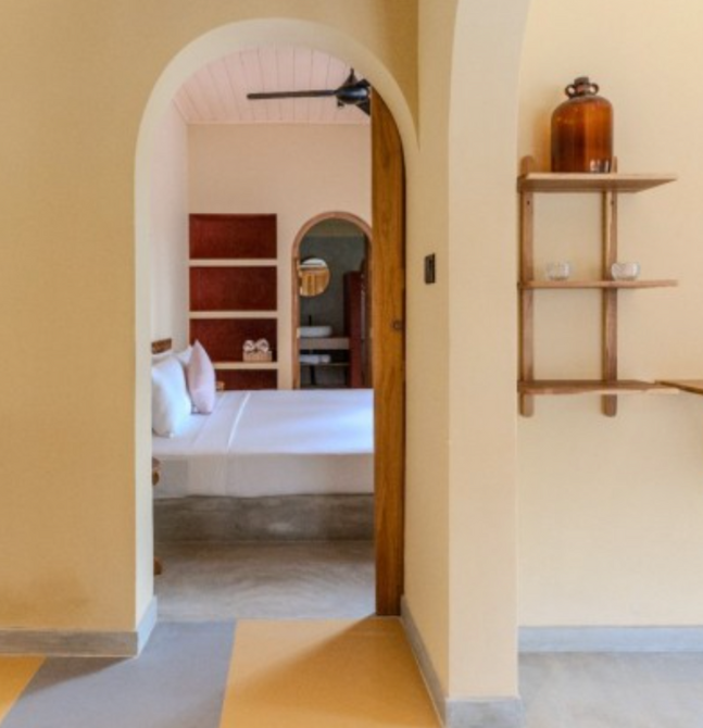Interior view of a stylish and minimalistic accommodation space with warm beige tones, arched doorway leading to a cozy bed with white linens, and a small wooden shelf with decorative items on the wall.