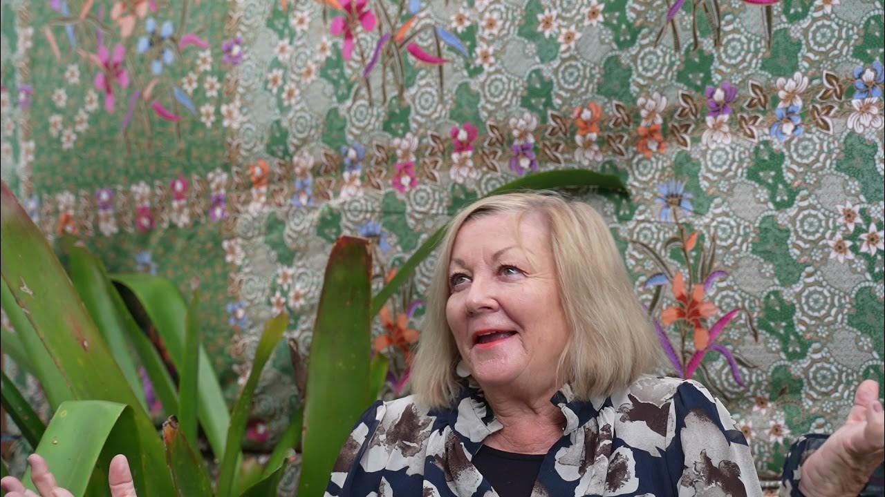 Suzie speaking during her yoga training testimonial, seated outdoors with tropical plants and a patterned green floral backdrop