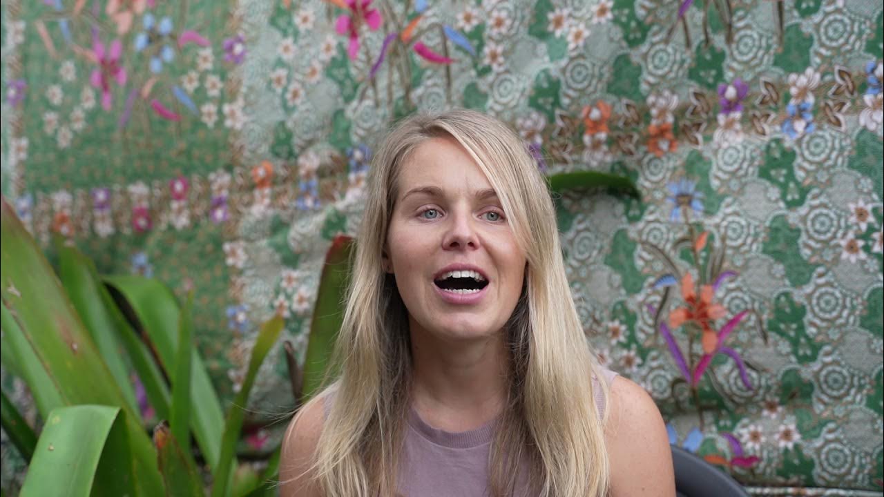 Tara sharing her experience during Santosha Yoga Teacher Training, seated in front of a colourful floral fabric backdrop with lush green plants