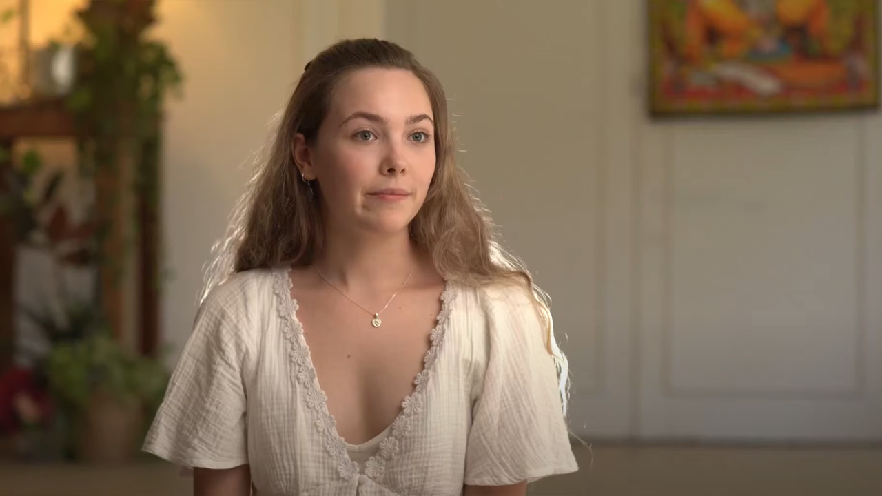 Young woman in a white blouse smiling softly while sitting indoors, speaking on camera with plants in the background.