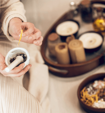 Hands holding small mortar and pestle while preparing herbal mixture beside tray of candles and oils